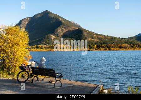 Tourist resting on wooden bench, relaxing at Vermilion Lakes lakeshore in autumn foliage season sunset time. Cycling in Banff National Park Stock Photo