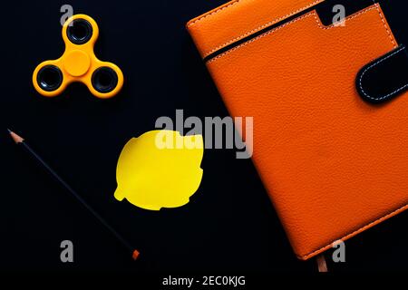 Orange notebook and spinner flat lay photo on black background. Fidget spinner and sticker note. Autumn season flat lay with art supplies. Hipster stu Stock Photo