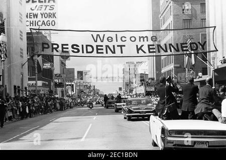 Inspection tour of NASA installations: Houston, Texas, motorcade, address at Rice University, 9:34AM. President John F. Kennedy (center right, in background) stands in the presidential limousine (Lincoln-Mercury Continental convertible) as his motorcade travels down Main Street in Houston, Texas, en route to Rice University. Photographers and members of the press ride in car in foreground; crowds line the street. Stock Photo