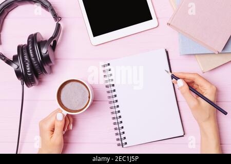 Workplace with blank notebook, female hands holding pencil and cup of coffee next to tablet PC, books and headphones, top view. Stock Photo