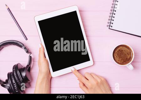 Female hands holding tablet with blank screen at workplace next to headphones, cup of coffee and notebook, flat lay. Stock Photo
