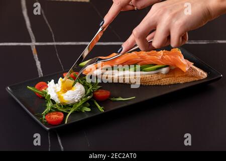hands cut Poached egg with toast, with smoked salmon, avocado, tomato and spinach. Stock Photo