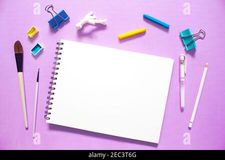 Artistic flat lay with white paper notepad, watercolor paints, brushes, and pastels, on violet background. Trendy workplace mockup for creative hobby. Stock Photo