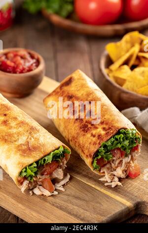 Chicken burritos with lettuce and tomato on a wooden cutting board with tortilla chips and salsa in background Stock Photo