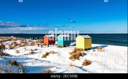 FINDHORN BAY MORAY FIRTH SCOTLAND WALKERS AND THREE COLOURFUL HUTS OR CHALETS  ON A SNOW COVERED BEACH IN WINTER Stock Photo