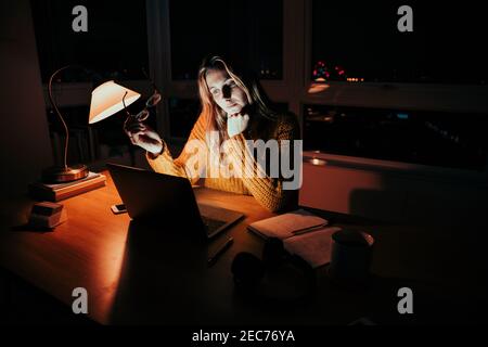 Caucasian business woman holding spectacles working late at night reading online article in home office Stock Photo