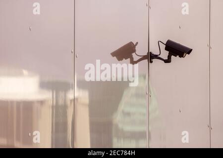 CCTV camera on toned glass wall of a building with reflection of modern city buildings. Security system and monitoring concept. Copy space. Stock Photo