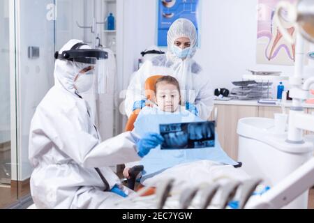 Orthodontist in ppe suit using dental radiography to explain dentistiry procedure for mouth hygine. Stomatolog in protectie suit for coroanvirus as safety precaution holding child teeth x-ray during consultation. Stock Photo