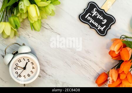 Spring Forward Daylight Saving Time concept with text on marble flat lay.  White alarm clock with tulips. Stock Photo