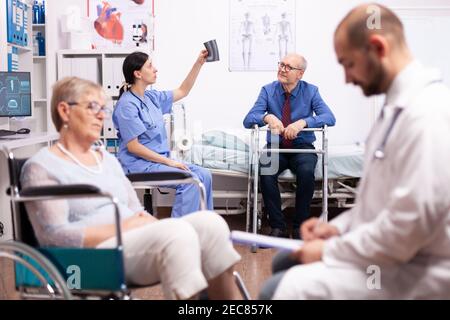 Nurse looking at radiography of handicapped patient sitting on wheelchair. Man with disabilities ,walking frame sitting in hospital bed. Health care system, clinic patients. Stock Photo