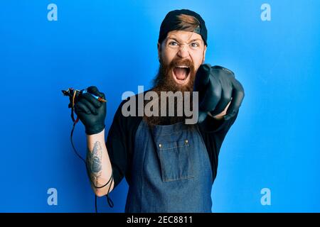 Redhead man with long beard tattoo artist wearing professional uniform and gloves pointing displeased and frustrated to the camera, angry and furious Stock Photo