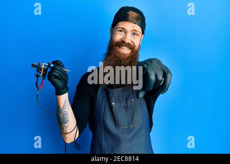 Redhead man with long beard tattoo artist wearing professional uniform and gloves pointing to you and the camera with fingers, smiling positive and ch Stock Photo