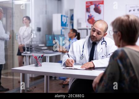 Specialist doctor prescribing treatment for senior woman writting on clipboard. Older patient visiting physician telling about health complaints, medical practitioner asking questions talking about filling disease. Stock Photo