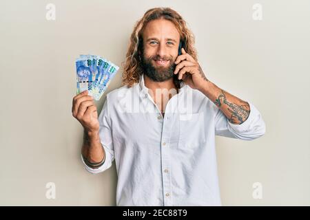Handsome man with beard and long hair talking on the phone holding 100 brazilian reals smiling with a happy and cool smile on face. showing teeth. Stock Photo