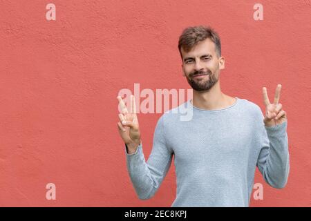 Handsome young man in gray shirt showing victory fingers sign, peace sign or number two with both hands on orange wall background. Attractive bearded Stock Photo
