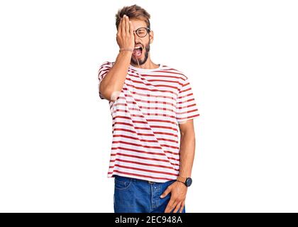 Handsome blond man with beard wearing casual clothes and glasses yawning tired covering half face, eye and mouth with hand. face hurts in pain. Stock Photo