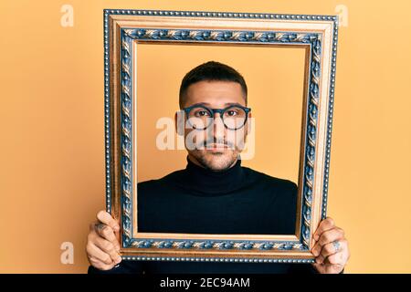 Handsome man with tattoos holding empty frame relaxed with serious expression on face. simple and natural looking at the camera. Stock Photo