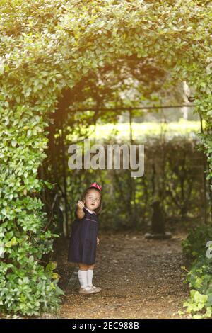 Beautiful preschool toddler aged little girl peeking out from a greenery covered arch Stock Photo