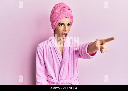 Young man wearing woman make up wearing shower towel on head and bathrobe pointing with finger surprised ahead, open mouth amazed expression, somethin Stock Photo