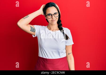 Young hispanic woman wearing professional waitress apron confuse and wonder about question. uncertain with doubt, thinking with hand on head. pensive Stock Photo