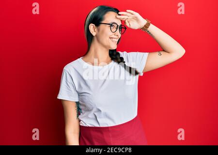 Young hispanic woman wearing professional waitress apron very happy and smiling looking far away with hand over head. searching concept. Stock Photo