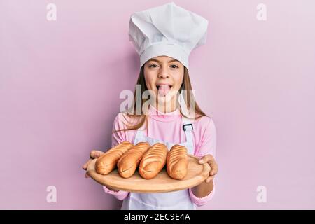 Beautiful brunette little girl wearing baker uniform holding homemade bread sticking tongue out happy with funny expression. Stock Photo