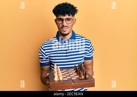 Young arab handsome man holding chess board wearing glasses clueless and confused expression. doubt concept. Stock Photo