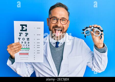 Handsome middle age man holding optometry glasses and medical exam sticking tongue out happy with funny expression. Stock Photo