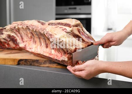 Chef is preparing prime rib steaks from a large prime beef rib roast with multiple bones. The meat is on a wooden board which is sitting on a modern kitchen counter. High quality photo Stock Photo