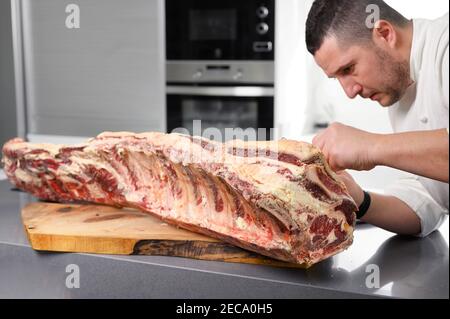 Chef is preparing prime rib steaks from a large prime beef rib roast with multiple bones. The meat is on a wooden board which is sitting on a modern kitchen counter. High quality photo Stock Photo