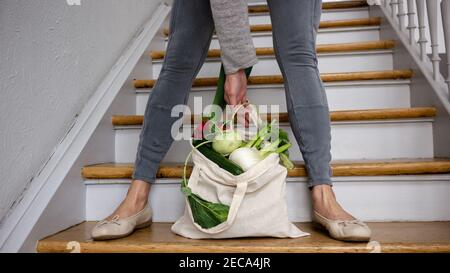 Woman in leisure outfit with a plastic-free groceries shopping bag full of local green vegetables on the wooden staircase: zero waste shopping.