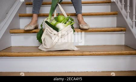 Woman in leisure outfit with a plastic-free shopping bag with local green vegetables on the wooden staircase: zero waste shopping. No plastic.