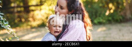Mother hugging pacifying sad upset crying toddler girl. Family young mom and crying baby in park outdoor. Bonding relationship of mom and child baby. Stock Photo