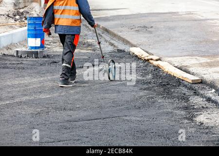 A road service engineer measures the work area with an electronic measuring wheel to order the right amount of fresh asphalt for paving. Stock Photo