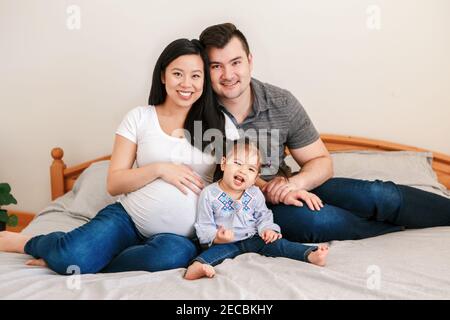 Family Asian Chinese pregnant woman and Caucasian man with mixed race toddler girl sitting on bed at home. Mother, father, baby daughter expecting new Stock Photo