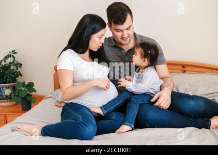 Family Asian Chinese pregnant woman and Caucasian man with toddler girl sitting on bed at home. Mother and father talking explaining to baby daughter Stock Photo