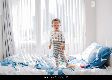 Cute little boy toddler standing on bed in room at home and looking at camera. Adorable innocent baby child in nursery room staring watching. Lonely k Stock Photo