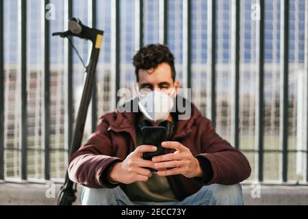 Handsome guy with face mask sending a message with his cell phone sitting on his electric scooter. Focus on the phone. Stock Photo