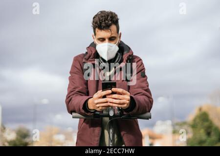 Handsome guy with face mask sending a message with his cell phone on the electric scooter. Stock Photo
