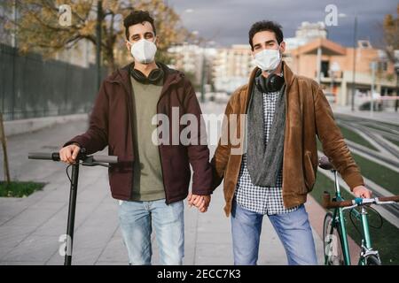 Two gay guys holding hands while walking with a bicycle and an electric scooter looking at the camera. Coronavirus concept. Stock Photo