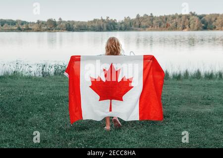 Girl wrapped in large Canadian flag by Muskoka lake in nature. Canada Day celebration outdoor. Kid in large Canadian flag celebrating national Canada Stock Photo