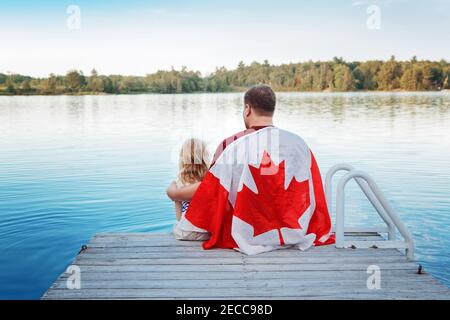Father and daughter wrapped in large Canadian flag sitting on wooden pier by lake. Canada Day celebration outdoor. Dad and child sitting together on 1 Stock Photo
