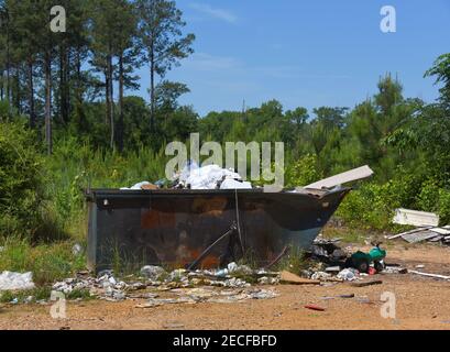Green dumpster on a county road is littered with overflowing trash.  Toy, siding, newspapers and plastic bottles cover the ground around the dumpster. Stock Photo