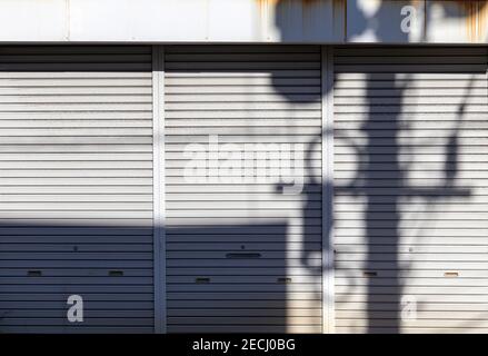 An electricity pole, its cables and transmission box, cast on odd shadow on the metal shutters of a closed shop and lends itself thus for grungy backg Stock Photo