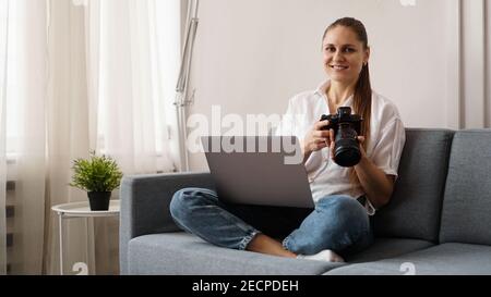 Happy young woman with photo camera using laptop at home. The photographer is holding the camera and smiling. Stock Photo