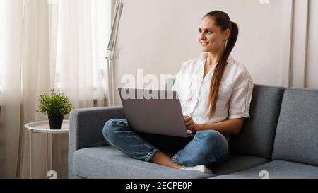Smiling happy woman sitting on the sofa and using laptop. The concept of remote work or study during quarantine. Stock Photo