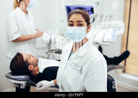 Woman working in dental clinic with patient sitting in chair and doctor in background. Dentist's assistant wearing face mask looking at camera. Stock Photo