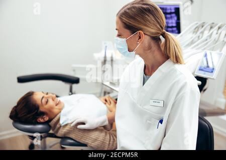 Female dentist wearing face mask talking with patient in dentist's chair. Dental doctor with female patient visiting for routine checkup. Stock Photo
