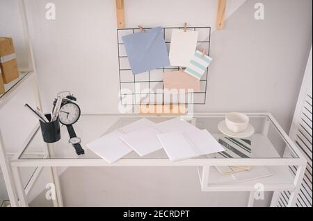 Background image of empty home cozy office working space modern scandinavian design. White glass table, holder lattice for notes with stylish decor elements, stationery, alarm clock and wristwatch Stock Photo
