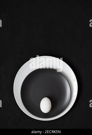 A white egg on a black plate on a black background. Black Easter concept. Vertical orientation. Minimalism. View from above. Space for text. Stock Photo